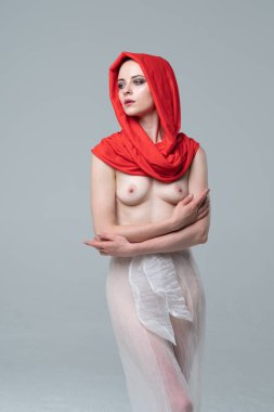 Stüdyoda çıplak poz veren genç ve güzel bir kız, kırmızı atkı ve beyaz pantolonla duruyor.