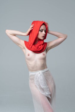 Stüdyoda çıplak poz veren genç ve güzel bir kız, kırmızı atkı ve beyaz pantolonla duruyor.