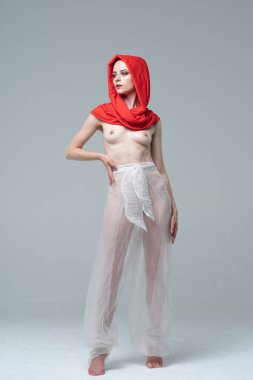 Stüdyoda çıplak poz veren genç ve güzel bir kız, kırmızı atkı ve beyaz pantolonla duruyor.