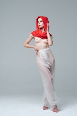 Stüdyoda çıplak poz veren genç ve güzel bir kız, kırmızı atkı ve beyaz pantolonla duruyor.