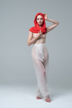 Stüdyoda çıplak poz veren genç ve güzel bir kız, kırmızı atkı ve beyaz pantolonla duruyor.