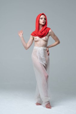 Stüdyoda çıplak poz veren genç ve güzel bir kız, kırmızı atkı ve beyaz pantolonla duruyor.