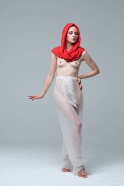 Stüdyoda çıplak poz veren genç ve güzel bir kız, kırmızı atkı ve beyaz pantolonla duruyor.