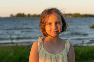 küçük bir kız portresi sarı bir elbise içinde bir göl yakınında duruyor