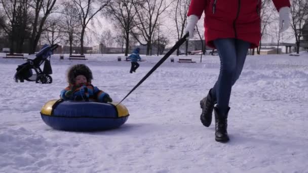 Bonito pequeno bebê mãe rola em tubulação na neve em um dia ensolarado, câmera lenta — Vídeo de Stock