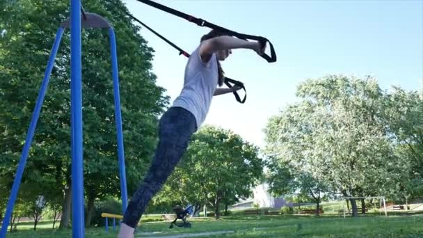 Joven bonita mujer haciendo ejercicios push-up en bucles de fitness en un parque. 4K lento mo desde abajo metraje — Vídeo de stock