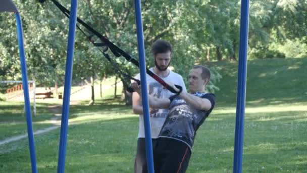 Vousatý fitness trenér řídí mladý muž intenzivní dělá shybů na fitness smyčky v letním parku. 4 k pomalé mo záběry — Stock video