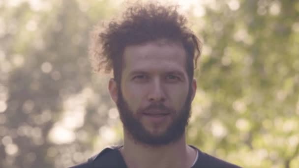 Homme barbu charismatique regardant à la caméra, puis souriant dans le parc d'été ensoleillé. 4K vue de face slowmo — Video