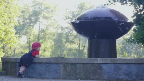 Baby with interest looking at the water standing near the fountain in the shadow of summer park. 4k slow mo footage — Stock Video