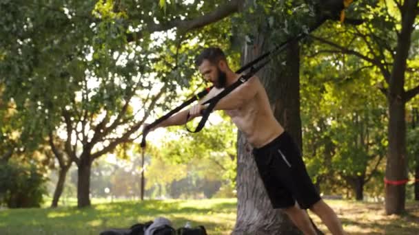 Бородатый молодой мускулистый мужчина начинает отжиматься с фитнес-петлями в солнечном парке. Съемка замедленного видео в 4К — стоковое видео