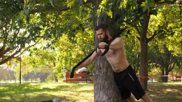 Бородатый молодой спортсмен делает отжимания тренировки упражнения с фитнес-петель возле дерева. Отснятый крупным планом снимок замедленной съемки 4К — стоковое видео