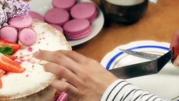 Manos de las mujeres poniendo en un plato un pedazo de pastel decorado con macarrones, fresas en rodajas y flores de color lila, 4K lento mo — Vídeo de stock