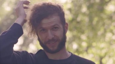 Genç adam kamera bakan sakallı ve darmadağınık saç sunshining Park düzeltiyor. 4 k ön görünüm slowmo