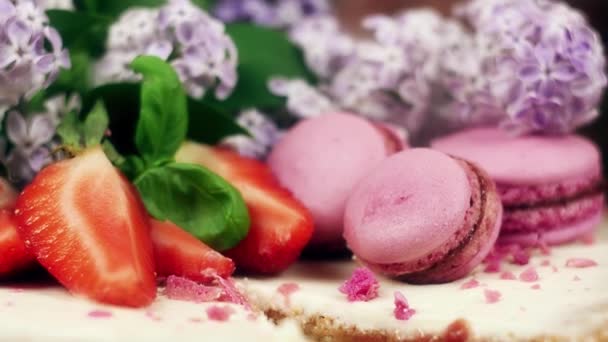 Kuchen mit einem Ausschnitt dekoriert mit Makronen, geschnittenen Erdbeeren und Fliederblüten, 4k slow mo — Stockvideo