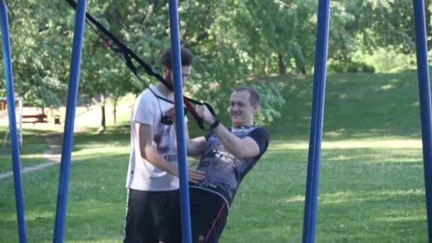 Joven comienza a hacer pull-ups en bucles de fitness bajo el control de entrenador de fitness barbudo en el parque de verano. 4K lento mo metraje — Vídeo de stock