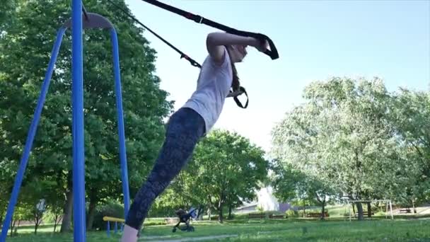 Mujer joven haciendo flexiones en trx fitness en el parque. 4K lento mo desde abajo metraje — Vídeos de Stock