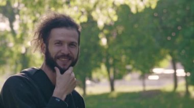 Sakalını yaz park, gülümseyen ve düzgünleştirir parlayan Güneşte ayakta bir genç sakallı adam portresi. 4 k yarı açık yan görünümü slowmo
