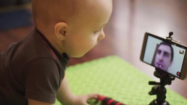 Niño pequeño sentado en el suelo es skyping en videollamada en un teléfono inteligente en trípode con su padre y luego cae trípode. 4K lento mo — Vídeos de Stock