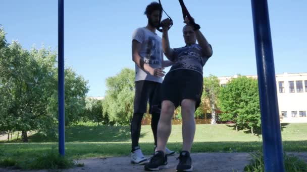 Człowiek zaczyna robić pull-up na fitness pętli pod kontrolą trenera fitness Brodaty w parku. 4 k zwolnionym tempie od dołu materiału — Wideo stockowe