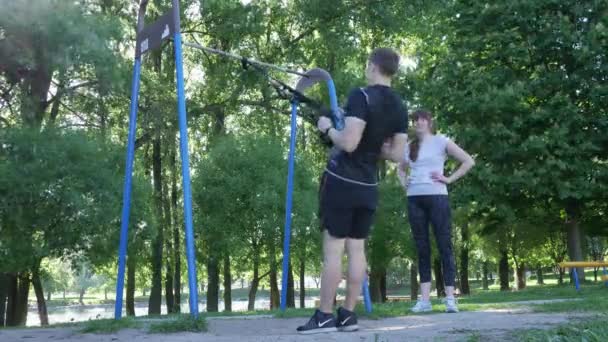 Ung kvinna som tittar mannen som gör pull-up exercerande på gym loopar i sommaren park. 4 k slow motion film — Stockvideo