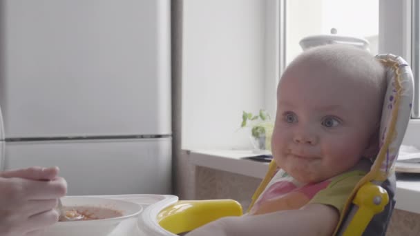 Mooie kleine babyjongen voeden met een lepel op de tafel door zijn moeder en kijken in de camera close-up weergave langzame mo video in 4k — Stockvideo