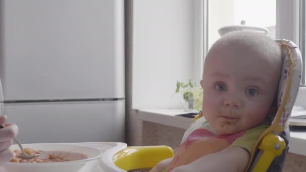 Een mooie kleine babyjongen voeden met een lepel aan de tafel terwijl hij kijken terug en dan kijken naar de camera en erachter op zijn vader moeder close-up weergave langzame mo video in 4k — Stockvideo