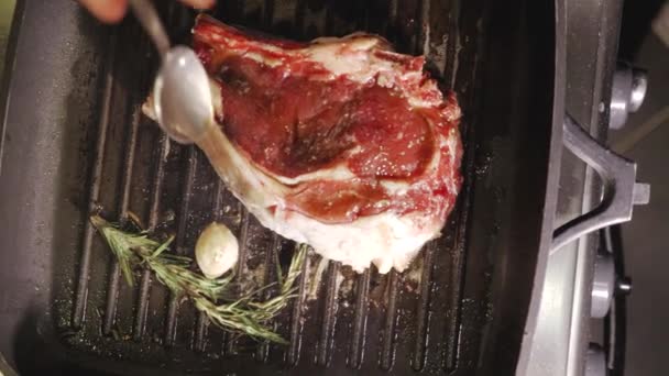 Unrecognizable man chefs hand pouring oil on a fresh beef steak wich roasting with rosemary and garlic on a hot black pan close up view from above slow motion video in 4K — Stock Video