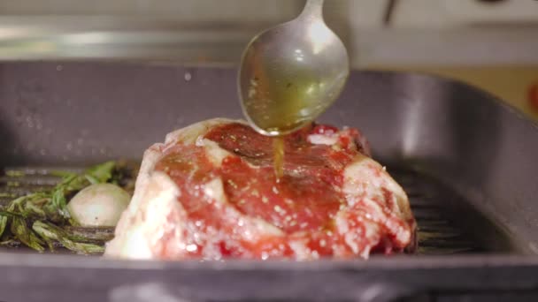 Un steak de bœuf rouge cru rôtissant sur une poêle à griller chaude noire et verser de l'huile dessus par cuillère dans la main des chefs de l'homme méconnaissable, vue rapprochée vidéo au ralenti en 4K — Video