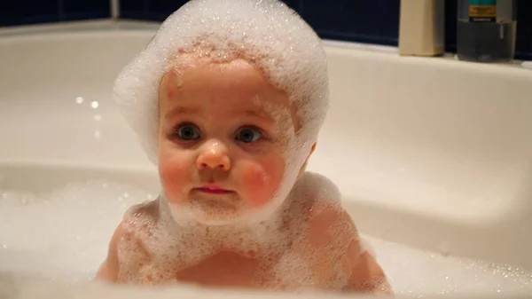 Младенец, покрытый пузырьками крупным планом, играет и улыбается в ванной. Ребенок с пеной на голове купается в ванне — стоковое фото