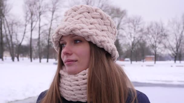 Beyaz örme şapka ve eşarp ayakta kamera bakıyor kış şehir parkında genç güzel bir kadının portresi soldan sağa 4k yavaş hareket video yakın sola çevirerek. — Stok video