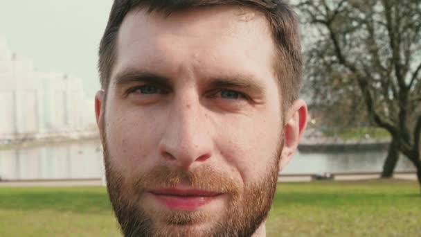 Портрет бородатого человека, который смотрит в камеру, а затем улыбается и смотрит в сторону на фоне летнего города замедленной съемки крупным планом видео в 4K — стоковое видео
