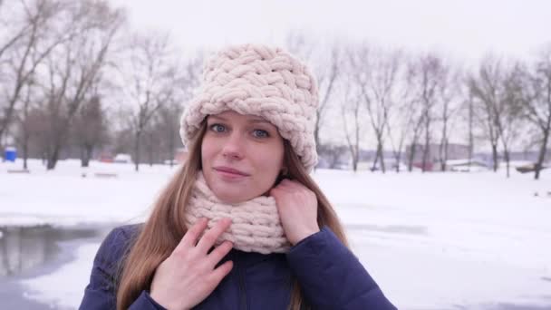 Porträt einer jungen hübschen Frau im winterlichen Stadtpark, die in die Kamera blickt, während sie ihre langen Haare und ihre weiße Strickmütze und ihren Schal in 4k Zeitlupe korrigiert. — Stockvideo