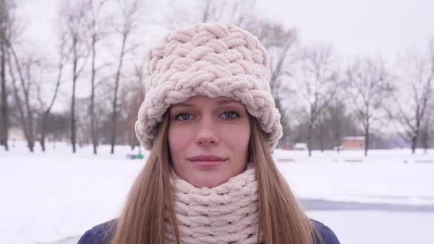 Retrato de uma mulher bonita de olhos azuis em chapéu de malha branca e cachecol em pé no parque da cidade de inverno e olhando para a câmera, em seguida, vira a cabeça em câmera lenta 4K fechar vídeo . — Vídeo de Stock