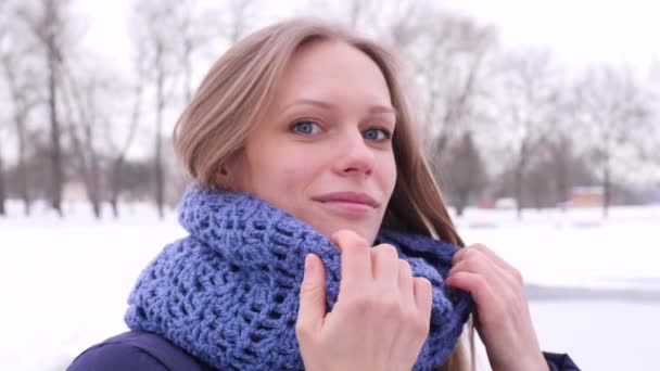 Egy csinos kék szemű nő portréja a téli városi parkból, aki a kamerába néz, és kijavítja hosszú haját és kék kötött sálját 4K lassított felvételen. — Stock videók