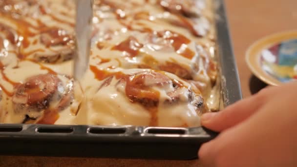 Woman or mans hands cuts with a special knife from a dish a fresh hot cinnamon bun spreaded with a white creamy-caramel cream and put in on the beautiful plate in slow motion 4K video. — Αρχείο Βίντεο
