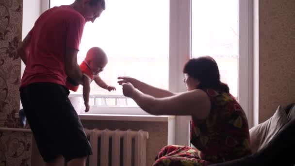 Senior man och kvinna leker med sin söta lilla barnbarn nära fönstret, och farfar skakar pojken i famnen medan mormor försöker fånga ungen med händerna i slow motion — Stockvideo