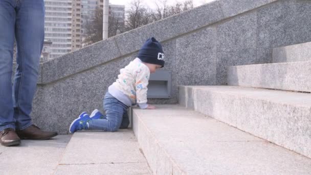 Bebé niño está subiendo las escaleras de granito en el parque de la ciudad y su padre pone al niño en sus pies y le ayudan en el fondo soleado día de primavera en cámara lenta toma 4K video — Vídeo de stock