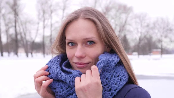 Porträtt av en vacker blåögd kvinna i vinterstadsparken som tittar på kameran och korrigerar sitt långa hår och blå stickade halsduk i 4K slow motion närbild video. — Stockfoto