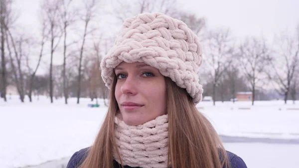 Portrait of a blue-eyed beautiful woman in white knitted hat and scarf standing in the winter city park and looking at the camera then turns her head in 4K slow motion close up video.