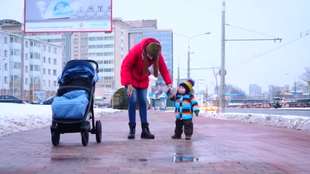 Młoda kobieta w czerwonej kurtce w dół prowadzi jej synka ręka spacerując po zimowej ulicy miasta w zwolnionym tempie średnio nakręcony 4k wideo. — Wideo stockowe