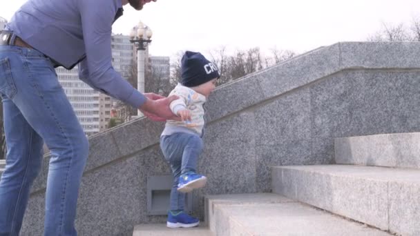 Hombre barbudo en gafas de sol sosteniendo a su pequeño hijo por la mano ayuda al niño a subir las escaleras en el parque de la ciudad en el fondo soleado día de primavera en cámara lenta toma 4K video — Vídeos de Stock