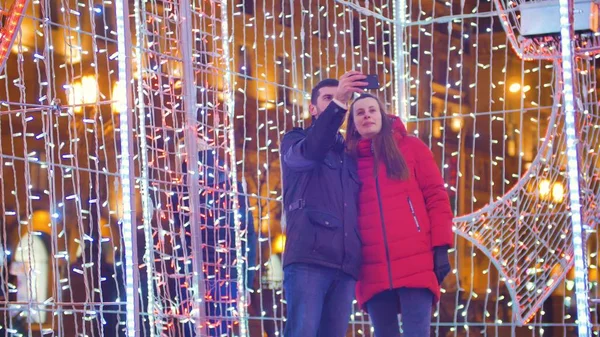 Nuori hauska pari tekee selfie yöllä kaupungin taustalla kirkas värikäs seppeleitä lahjapakkauksessa — kuvapankkivalokuva