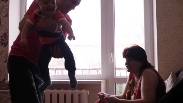 Senior homme et femme jouent avec leur mignon petit-fils près de la fenêtre, et grand-père secoue le garçon dans ses bras tandis que grand-mère effrayer l'enfant avec ses mains et il sourit lentement — Video