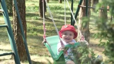 yüzünde sivrisinek ısırıkları ile kırmızı panama mutlu gülümseyen küçük bebek arka planda ağaçlar ile parkta bir salıncak üzerinde sürme ve daha sonra annesi yavaş hareket yan görünümü 4k video ona geliyor.