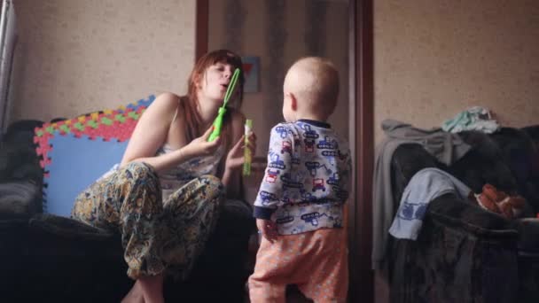 Een jonge mooie vrouw blazen zeepbellen en haar kleine jongen speelt vangen ze gelukkig glimlachen in de kamer in slow motion 4K video — Stockvideo