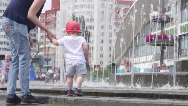 Young woman holding her baby son hand near the fountain and showing her boy the falling water with the city center on background. 4k slow motion back view video. — 비디오
