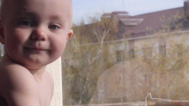 Küçük bebek çocuk yakın pencerenin yanında oturuyor, içine bakmak, sonra kamera içine ve yavaş hareket 4k pencerenin arkasında bir ağacın arka planda pencerenin dışında bir şey işaret