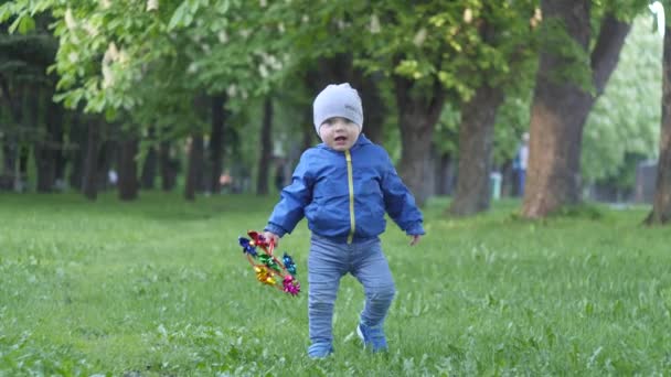 Kleine schattige baby jongen in een blauwe jas met een speelgoed in de hand gaat op het gazon naar de camera in het stadspark met bomen op de achtergrond in slow motion 4k-video — Stockvideo