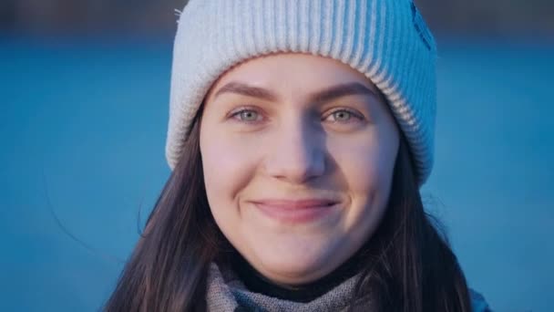美しい緑の目のブルネットの女の子の女性の肖像画誰がカメラを見ています,魅力的な笑い,点滅し、その後、彼女の白いニット帽を修正します湖とともにバックグラウンドでクローズアップ4kビデオ. — ストック動画