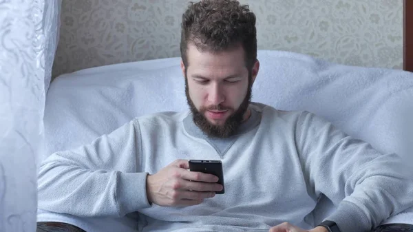 Young man talking on cellphone and drinking coffee on chair at home — Stock Photo, Image